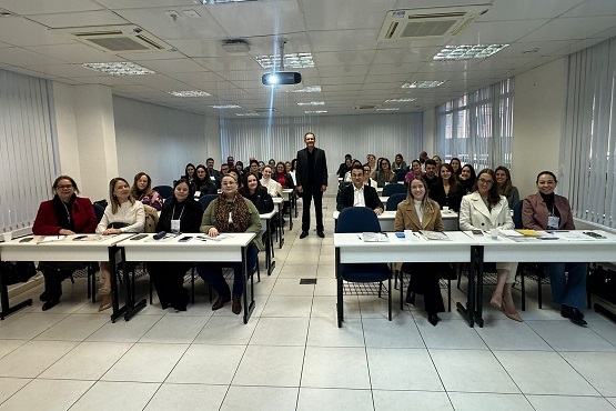 A imagem mostra um grupo de pessoas sentadas em torno de uma mesa em uma sala de aula ou reunião. A mesa é em formato de "U" e as pessoas estão olhando para a câmera. Há cerca de 20 pessoas na foto, a maioria delas está sorrindo. A sala tem piso de cerâmica clara e paredes brancas. No fundo, há uma tela de projeção e um projetor no teto. A iluminação é feita por lâmpadas fluorescentes.