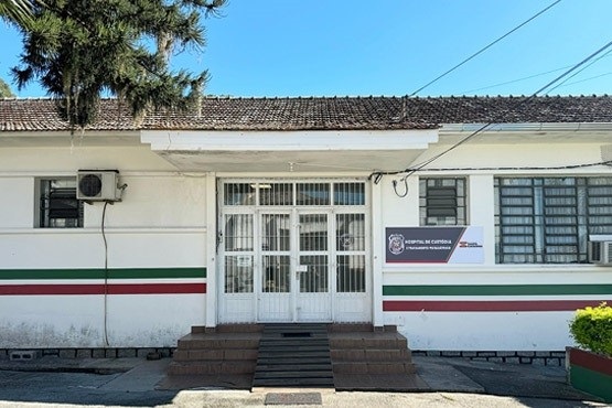 A imagem mostra a fachada de um prédio com uma escadaria central que leva a uma porta de vidro. O prédio tem faixas horizontais nas cores verde, branca e vermelha. Há uma placa ao lado da porta com o logotipo e o nome de uma instituição, mas o texto não está totalmente legível na imagem. No lado direito da imagem, há uma árvore alta e um céu azul claro.