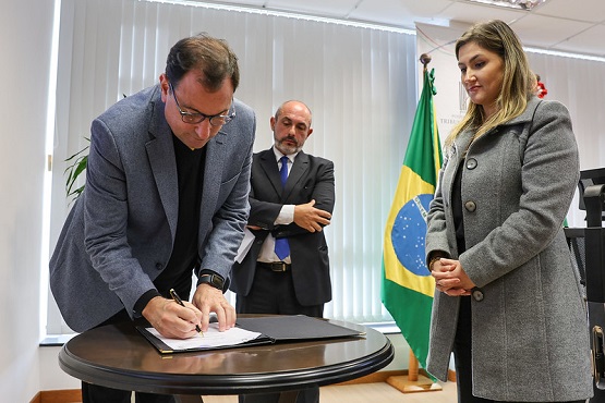A imagem mostra três pessoas em um ambiente formal. Um homem de terno cinza está assinando um documento sobre uma mesa redonda. Atrás dele, há outro homem de terno escuro e gravata azul, observando a assinatura. À direita, uma mulher de casaco cinza está de pé, com as mãos cruzadas na frente do corpo, olhando para a cena. Ao fundo, há uma bandeira do Brasil e outra bandeira verde.
