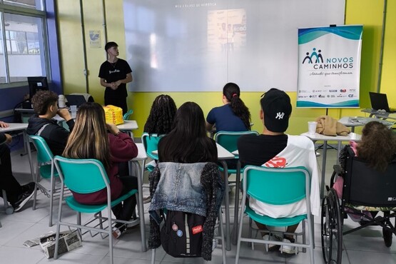 A imagem mostra uma sala de aula com várias pessoas sentadas em cadeiras verdes, voltadas para a frente da sala, onde um homem está de pé, aparentemente dando uma palestra ou aula. Na parede ao fundo, há um banner com o texto "Novos Caminhos" e alguns logotipos. As paredes da sala são amarelas e há uma janela à direita. Algumas pessoas estão usando máscaras, e uma pessoa em uma cadeira de rodas está presente na parte inferior da imagem.