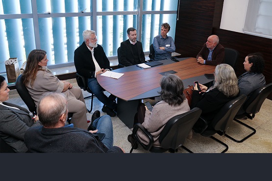 A imagem mostra uma reunião em uma sala de conferências. Há várias pessoas sentadas ao redor de uma mesa retangular de madeira. A maioria das pessoas está vestida formalmente, com ternos e roupas de trabalho. A sala tem grandes janelas ao fundo, permitindo a entrada de bastante luz natural.