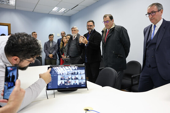 A imagem mostra um grupo de pessoas em uma sala de reunião. No centro da imagem, há uma mesa com um monitor de computador exibindo uma videoconferência. Algumas pessoas estão em pé ao redor da mesa, enquanto outras estão sentadas. Um homem de barba e óculos está tirando uma foto com seu celular. Ao fundo, há várias pessoas em pé, algumas delas usando terno e gravata. A sala tem um teto com iluminação embutida e paredes claras.