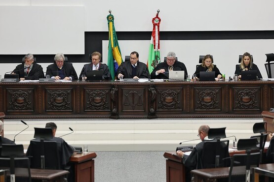 A imagem mostra uma sessão de um tribunal. Há várias pessoas sentadas em uma bancada elevada, provavelmente juízes ou desembargadores, todos vestidos com togas pretas. A bancada é de madeira escura e trabalhada, com detalhes ornamentais. Atrás da bancada, há bandeiras do Brasil e de um estado brasileiro, possivelmente Santa Catarina.