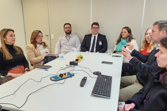 A imagem mostra uma reunião em uma sala de conferências. Há sete pessoas sentadas ao redor de uma mesa branca. Na mesa, há um teclado, um mouse, um controle remoto e alguns cabos. As pessoas estão vestidas formalmente, com ternos e roupas sociais. Algumas delas estão olhando para documentos ou dispositivos eletrônicos. No fundo, há uma parede clara.