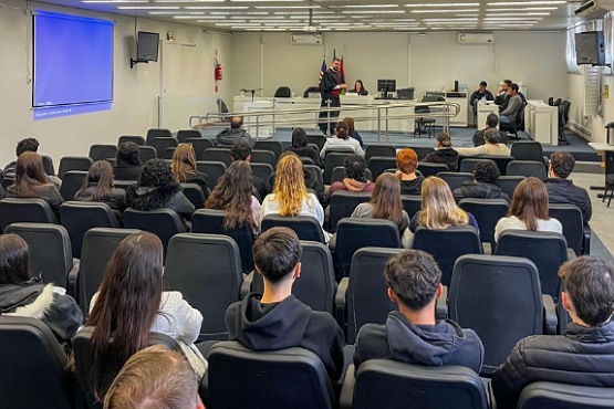 A imagem mostra uma sala de aula ou auditório com várias pessoas sentadas em cadeiras voltadas para a frente. A maioria das pessoas está de costas para a câmera. Na frente da sala, há uma pessoa em pé, aparentemente falando ou apresentando algo. Há também uma tela grande na parede da frente, que está acesa com uma cor azul. As cadeiras são de cor escura e estão dispostas em fileiras.