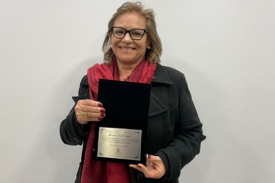 A imagem mostra uma mulher sorridente segurando uma placa de homenagem. Ela tem cabelos loiros e usa óculos. Está vestindo um casaco preto e um cachecol vermelho. A placa que ela segura é retangular e parece ser de metal, com uma inscrição que não é legível na imagem. O fundo da imagem é uma parede branca.