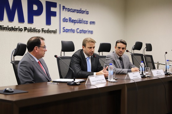 A imagem mostra três homens sentados atrás de uma mesa em uma sala de conferências. Eles estão participando de uma coletiva de imprensa ou reunião formal. Na parede atrás deles, há um grande logotipo com as letras "MPF" em azul, que significa Ministério Público Federal. 	Ao lado do logotipo, está escrito "Procuradoria da República em Santa Catarina".  Os três homens estão vestidos formalmente com ternos. O homem à esquerda está usando um terno cinza escuro e gravata vermelha. O homem no centro está falando ao microfone e gesticulando com as mãos; ele está usando um terno preto e gravata azul clara. O homem à direita está usando um terno cinza claro e gravata azul escura.  Na mesa, há microfones, garrafas de água e placas de identificação na frente de cada homem. A sala parece ser um ambiente formal e profissional.