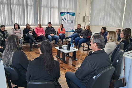 A imagem mostra um grupo de pessoas sentadas em cadeiras dispostas em círculo em uma sala. Há cerca de 18 pessoas, a maioria mulheres, participando de uma reunião ou discussão. No centro do círculo, há uma pequena mesa com alguns objetos em cima. Ao fundo, há uma janela grande com persianas brancas e um banner com o logotipo e o nome "Novos Caminhos". A sala tem piso de madeira e parece ser um ambiente de trabalho ou uma sala de reuniões.