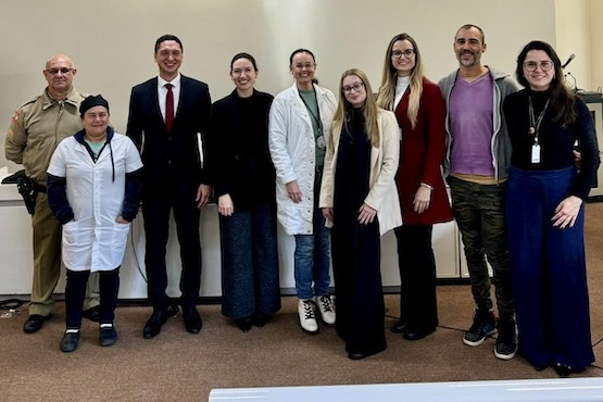 A imagem mostra um grupo de nove pessoas posando para uma foto em um ambiente interno. Da esquerda para a direita:  1. Um homem de uniforme bege, possivelmente um policial. 2. Uma mulher de uniforme branco, com um lenço preto na cabeça. 3. Um homem de terno preto e gravata vermelha, sorrindo. 4. Uma mulher de blusa preta e calça escura. 5. Uma mulher de jaleco branco e calça jeans, sorrindo. 6. Uma mulher de cabelo loiro, usando óculos, casaco claro e calça escura. 7. Uma mulher de cabelo loiro, usando óculos, casaco vermelho e calça escura. 8. Um homem de barba, usando uma blusa roxa e calça camuflada. 9. Uma mulher de cabelo escuro, usando óculos, blusa cinza e calça azul escura.  Todos estão de pé, lado a lado, e parecem estar em um ambiente formal, possivelmente uma sala de conferências ou um auditório.