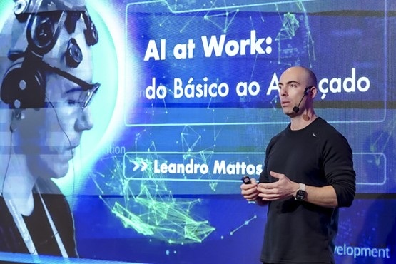 A imagem mostra um homem careca, usando uma camiseta preta de manga longa, falando em um palco. Ele está usando um microfone de cabeça e um relógio no pulso esquerdo. Atrás dele, há uma grande tela de projeção com a seguinte mensagem: "AI at Work: do Básico ao Avançado". Abaixo dessa frase, está o nome "Leandro Mattos". No lado esquerdo da tela, há uma imagem de perfil de uma pessoa usando um dispositivo na cabeça, possivelmente um equipamento de monitoramento cerebral. A tela de fundo tem um design tecnológico com linhas e pontos conectados, sugerindo um tema de inteligência artificial.