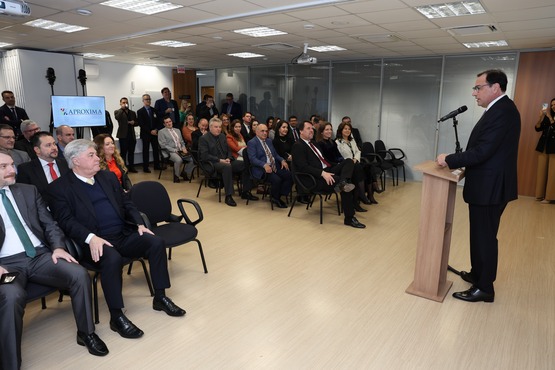 A imagem mostra uma sala de conferências com várias pessoas sentadas, prestando atenção a um homem que está falando em um púlpito com um microfone. O homem está localizado no lado direito da foto e usa terno escuro e óculos. As pessoas na plateia também estão vestidas formalmente, com ternos e roupas sociais. No fundo da sala, há uma placa com o logotipo "APROXIMA". A sala tem um piso de madeira clara e iluminação de teto. As paredes são de vidro e há algumas cadeiras vazias.