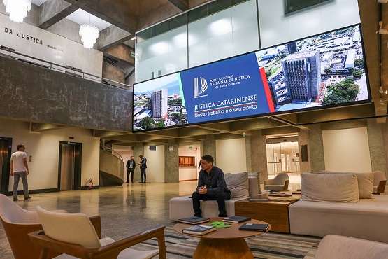 A imagem mostra o interior de um prédio moderno. No centro da imagem, há um homem sentado em um sofá branco, olhando para baixo. Ao redor dele, há outros sofás e mesas de centro com revistas e papéis.  No fundo, há um grande painel eletrônico com imagens de edifícios e o texto "Justiça Catarinense - Nosso trabalho é o seu direito".  À esquerda, há um elevador e algumas pessoas caminhando pelo local. O ambiente é bem iluminado e tem um design contemporâneo, com paredes de concreto e móveis modernos.