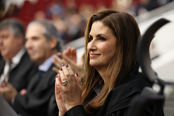 A imagem mostra uma mulher de cabelos castanhos longos e lisos, vestindo uma toga preta, aplaudindo. Ela está sorrindo e parece estar em um evento formal. Ao fundo, há outras pessoas também vestindo togas, desfocadas. A mulher usa anéis e esmalte escuro nas unhas.