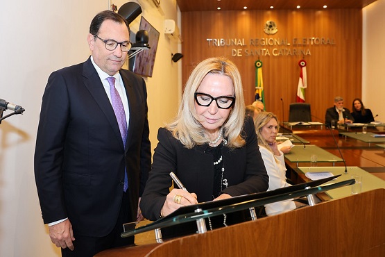 A imagem mostra uma mulher loira de óculos grandes e pretos assinando um documento em um púlpito. Ela está vestida com um blazer preto. Ao lado dela, há um homem de terno escuro, camisa branca e gravata roxa, observando a assinatura. Ambos estão em uma sala de tribunal, com um painel de madeira ao fundo onde se lê "Tribunal Regional Eleitoral de Santa Catarina". No fundo, há outras pessoas sentadas em uma mesa, incluindo uma mulher loira de cabelo curto e um homem de terno escuro. Há também bandeiras do Brasil e de Santa Catarina atrás deles.