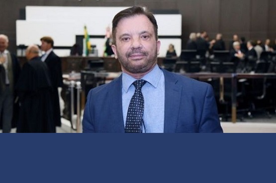 A imagem mostra um homem de meia-idade, com cabelo curto e barba bem aparada, vestindo um terno azul e uma gravata azul com bolinhas brancas. Ele está sorrindo e olhando diretamente para a câmera. Ao fundo, há um ambiente que parece ser uma sala de conferências ou um tribunal, com várias pessoas sentadas e uma bandeira do Brasil visível.
