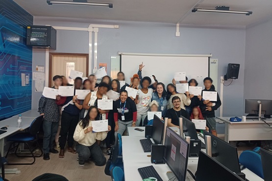 A imagem mostra um grupo de pessoas em uma sala de aula ou treinamento. Elas estão posando para a foto, algumas segurando papéis ou certificados. A sala tem mesas com computadores e cadeiras. Na parede ao fundo, há um quadro branco e um monitor de TV.