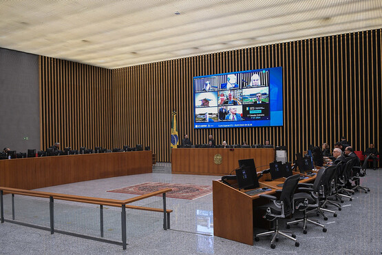 A imagem mostra uma sala de conferências moderna e espaçosa. No centro da imagem, há uma grande mesa em formato de "U" com várias cadeiras de escritório ao redor. Na parede ao fundo, há um grande painel de madeira com ripas verticais e uma tela grande exibindo várias janelas de videoconferência. A bandeira do Brasil está posicionada ao lado da tela. O chão é de mármore claro e há um tapete no centro da sala. A iluminação é suave e o ambiente parece ser formal, possivelmente uma sala de reuniões governamentais ou corporativas.