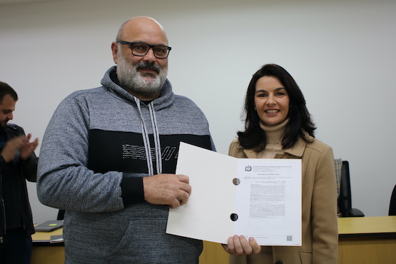 A imagem mostra duas pessoas em um ambiente interno. À esquerda, há um homem de meia-idade com barba grisalha, óculos e vestindo um moletom cinza com capuz. Ele está segurando um documento. À direita, há uma mulher de cabelos escuros e longos, vestindo um casaco bege. Ela também está segurando o documento junto com o homem. Ambos estão sorrindo. Ao fundo, há uma pessoa desfocada aplaudindo. O ambiente parece ser uma sala de reuniões ou um escritório, com uma mesa e cadeiras ao fundo.