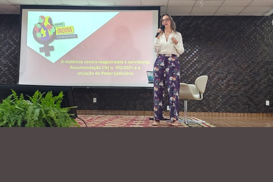 A imagem mostra uma mulher em pé, segurando um microfone, em um palco. Ela está vestindo uma blusa branca e calças com estampa floral. Atrás dela, há uma tela de projeção com um slide que contém o logotipo do "Programa Indira" e o texto: "A violência contra magistradas e servidoras, Recomendação CNJ n. 102/2021 e a atuação do Poder Judiciário". Ao lado da mulher, há uma cadeira vazia. Na frente do palco, há algumas plantas decorativas. O fundo da sala é composto por uma parede com revestimento escuro.
