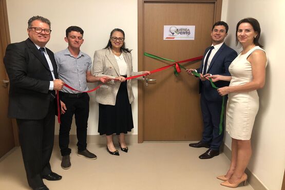 A imagem mostra um grupo de cinco pessoas participando de uma cerimônia de inauguração, onde estão cortando uma fita vermelha. Eles estão em frente a uma porta. A pessoa à esquerda está segurando a fita com uma tesoura, e as outras quatro pessoas estão ao lado, também segurando a fita. Todos estão bem vestidos, com roupas formais. No fundo, há uma placa na parede com o logotipo do Tribunal de Justiça de Santa Catarina (TJSC).