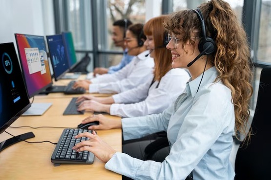 A imagem mostra várias pessoas sentadas em uma fileira, todas usando fones de ouvido com microfones e trabalhando em computadores. Elas estão em um ambiente de escritório, e cada uma parece estar digitando em um teclado. A imagem está rotacionada 90 graus para a esquerda.