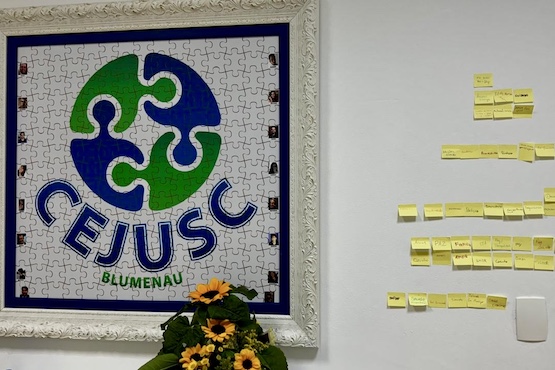 A imagem mostra uma parede, em sua parte superior, há um quadro com um quebra-cabeça montado que forma o logotipo do CEJUSC (Centro Judiciário de Solução de Conflitos e Cidadania) de Blumenau. O logotipo é composto por peças de quebra-cabeça em azul e verde que se encaixam, simbolizando a resolução de conflitos. No meio, há uma parede com vários post-its amarelos organizados em colunas. Parece ser um quadro de organização ou planejamento.