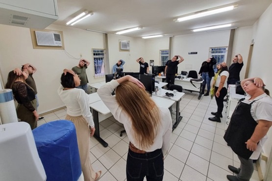 A imagem mostra um grupo de pessoas em uma sala, aparentemente participando de uma atividade de alongamento ou exercício. A sala tem piso de azulejo claro e paredes brancas. As pessoas estão em pé, espalhadas pela sala, com os braços levantados e mãos na cabeça, como se estivessem alongando o pescoço. Há mesas e cadeiras no ambiente, e algumas pessoas estão próximas a elas.