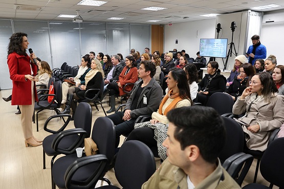 A imagem mostra uma sala de conferências com várias pessoas sentadas em cadeiras, todas voltadas para a frente, onde uma mulher de pé, vestida com um casaco vermelho, está falando ao microfone. A sala parece estar cheia, com pessoas de diferentes idades e estilos de roupa. No fundo, há uma tela de projeção e alguns equipamentos de gravação.