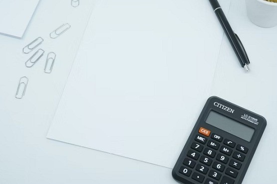 A imagem mostra uma calculadora preta da marca Citizen, um pedaço de papel em branco, um clipe de papel e uma caneta preta. A calculadora está posicionada no canto inferior esquerdo da imagem, a caneta está à direita e os clipes de papel estão no canto superior direito.