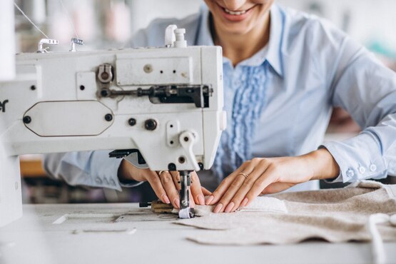 A imagem mostra uma pessoa utilizando uma máquina de costura. A pessoa está vestindo uma camisa azul clara e parece estar sorrindo enquanto trabalha. A máquina de costura é branca e ocupa a maior parte da imagem.