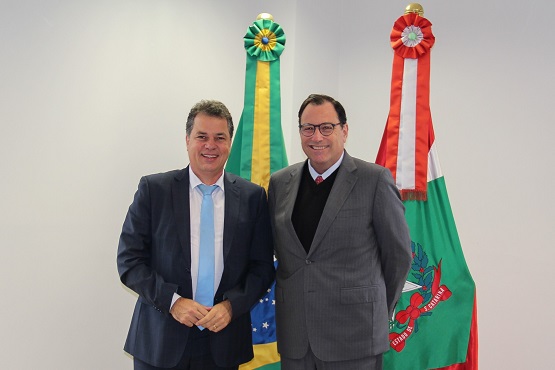 Foto horizontal: Francisco Oliveira Neto posa com autoridade entre bandeiras do Brasil e de SC, ambiente institucional, tons claros.