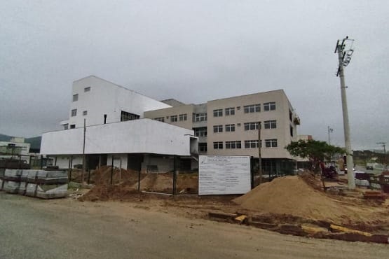 A imagem mostra um prédio em construção ou reforma, com andaimes e estruturas de suporte ao redor. O terreno em frente ao prédio parece estar em obras, com áreas de terra exposta e materiais de construção espalhados. O prédio é de cor clara e tem várias janelas.