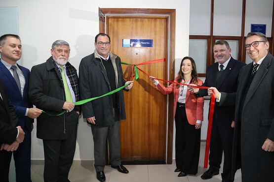 A imagem mostra um grupo de cinco pessoas em um evento de inauguração ou abertura. Eles estão em frente a uma porta de madeira com uma placa azul. Duas fitas, uma verde e uma vermelha, estão sendo cortadas. Todos parecem estar sorrindo e olhando para a câmera. A mulher está mais ao fundo, enquanto os homens estão mais à frente.