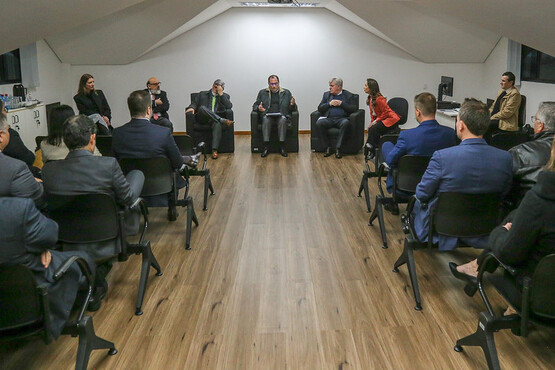 A imagem mostra uma reunião formal com várias pessoas sentadas em cadeiras dispostas em um círculo. A maioria dos participantes está vestida com trajes formais, como ternos e blazers. A sala tem um piso de madeira e parece ser bem iluminada. Há uma tela ou quadro branco na parede ao fundo.