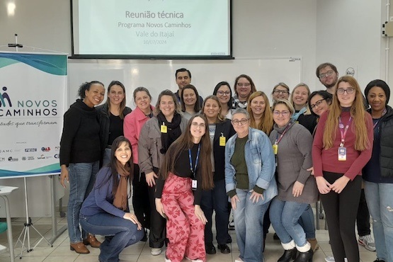 A imagem mostra um grupo de aproximadamente 20 pessoas, a maioria mulheres, posando para uma foto em um ambiente interno. Eles estão em frente a um quadro branco e um projetor que exibe a mensagem "Reunião técnica Programa Novos Caminhos Vale do Itajaí 10/07/2023". À esquerda, há um banner com o logotipo do programa "Novos Caminhos". As pessoas estão sorrindo e parecem estar em um ambiente de trabalho ou reunião. Algumas delas estão usando crachás.