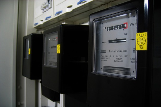 A imagem mostra três medidores de energia elétrica alinhados em uma superfície. Eles são de cor preta e possuem um visor transparente onde é possível ver os números que indicam a leitura do consumo de energia em kWh. Cada medidor tem um selo amarelo com informações técnicas. Ao fundo, há uma parede branca e parte de um painel elétrico com disjuntores.