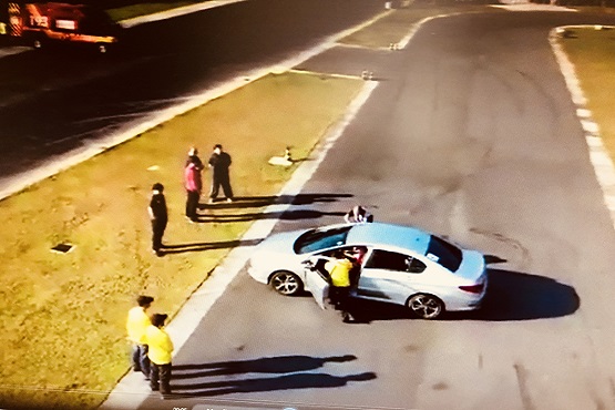 A imagem mostra uma cena ao ar livre, provavelmente em uma pista ou estacionamento. No centro da imagem, há um carro prata com as portas abertas. Perto do carro, há várias pessoas, algumas vestindo coletes amarelos. Parece que estão envolvidos em alguma atividade ou treinamento. Ao fundo, há mais pessoas observando, e à esquerda, pode-se ver um veículo de emergência, possivelmente um caminhão de bombeiros. A área ao redor é pavimentada, com uma faixa de grama ao lado.
