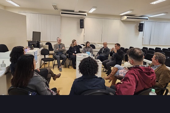 A imagem mostra um grupo de pessoas sentadas em círculo em uma sala de reuniões. Há um total de onze pessoas, todas sentadas em cadeiras. A sala tem paredes brancas, janelas com persianas brancas fechadas e iluminação fluorescente no teto. No fundo da sala, há um computador sobre uma mesa e algumas cadeiras vazias. As pessoas parecem estar em uma discussão ou reunião, e algumas delas estão gesticulando enquanto falam.