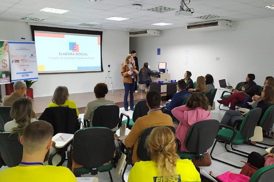 A imagem mostra uma sala de aula ou auditório com várias pessoas sentadas em cadeiras, voltadas para a frente, onde há uma apresentação sendo exibida em um projetor. As pessoas parecem estar assistindo a uma palestra ou treinamento. Algumas pessoas estão vestindo camisetas amarelas, enquanto outras estão com roupas de cores variadas. Na frente da sala, há uma tela de projeção com slides visíveis, mas o conteúdo dos slides não está legível na imagem. Ao lado da tela, há uma pessoa em pé, possivelmente o palestrante, e outra pessoa ao lado dela. A sala é bem iluminada e tem um ambiente de aprendizado ou reunião.