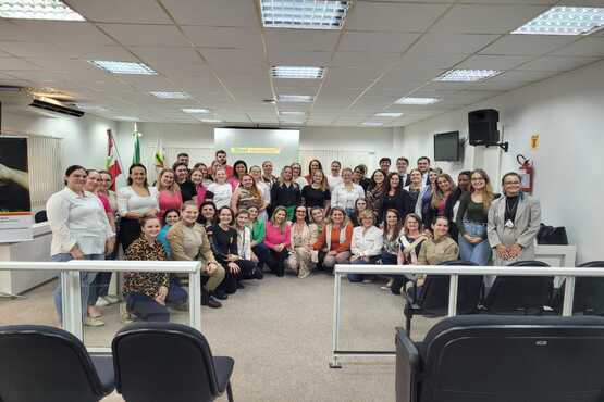A imagem mostra um grupo grande de pessoas posando para uma foto em um ambiente interno, que parece ser uma sala de conferências ou uma sala de reuniões. As pessoas estão organizadas em várias fileiras, algumas sentadas e outras em pé. Há uma mistura de homens e mulheres, e todos parecem estar sorrindo para a câmera. No fundo, há bandeiras e uma tela de projeção, sugerindo que o local pode ser uma instituição oficial ou um local de trabalho. O teto tem iluminação fluorescente e há cadeiras vazias na frente do grupo.