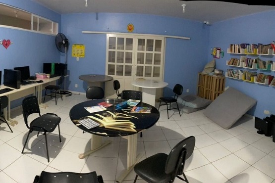 A imagem mostra uma biblioteca. As paredes são pintadas de azul claro e o piso é de cerâmica branca. No lado esquerdo da imagem, há uma mesa com dois computadores e cadeiras pretas. Acima da mesa, há uma janela com vidros transparentes. No centro da sala, há duas mesas redondas com cadeiras ao redor. Em cima de uma das mesas, há vários livros e materiais de estudo. No fundo da sala, há uma porta de vidro com cortinas brancas. À direita da porta, há uma estante de madeira com vários livros organizados. Perto da estante, há um colchão cinza dobrado e alguns travesseiros. A sala parece bem iluminada e organizada, com um ambiente propício para estudo ou atividades educacionais.