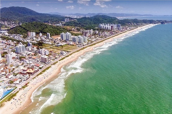 A imagem mostra uma vista aérea de uma cidade litorânea. À esquerda, há uma extensa faixa de mar verde, com ondas quebrando na praia. A praia tem areia clara e parece ser bastante larga. À direita da praia, há uma área urbana com muitos prédios altos e algumas áreas verdes. Mais ao fundo, é possível ver colinas ou montanhas cobertas de vegetação. A imagem está inclinada, com o mar na parte inferior e a cidade na parte superior.