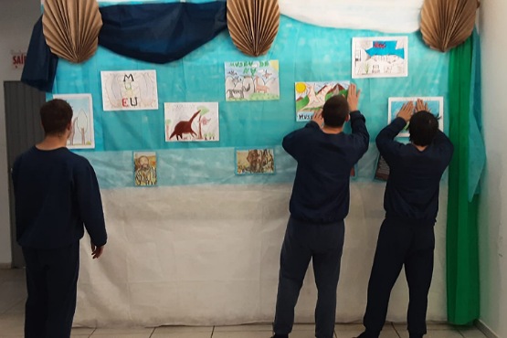 A imagem mostra três jovens de costas, todas vestindo roupas escuras, provavelmente uniformes. Elas estão em frente a um mural decorado com um tecido TNT azul e branco, com alguns detalhes em verde. No mural, há vários desenhos coloridos pendurados, que parecem ser feitos por crianças. Os desenhos incluem imagens de dinossauros, árvores, e outras figuras. As duas pessoas à direita estão ajustando ou colocando os desenhos no mural, enquanto a pessoa à esquerda está apenas olhando. O ambiente parece ser uma sala fechada.