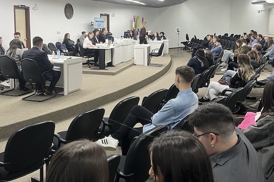 A imagem mostra uma sala de júri de um fórum. Há várias pessoas sentadas em cadeiras dispostas em fileiras, assistindo a uma sessão. Na parte da frente, há uma mesa comprida com várias pessoas sentadas, possivelmente autoridades ou membros de um conselho. Atrás dessas pessoas, há bandeiras de diferentes cores, provavelmente representando estados ou municípios. A sala é bem iluminada e tem um ambiente formal.