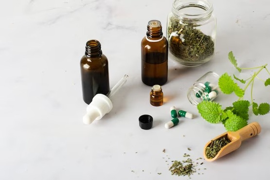 A imagem mostra uma variedade de itens relacionados a ervas e medicamentos naturais sobre uma superfície branca. Há dois frascos de vidro âmbar, um maior e outro menor, ambos com conta-gotas. Ao lado deles, há um pequeno frasco de vidro transparente com uma tampa de metal, contendo cápsulas verdes e brancas. Também há um pote de vidro com tampa de metal, cheio de ervas secas. Perto do pote, há uma colher de madeira com mais ervas secas. À direita, há um ramo de folhas verdes frescas.
