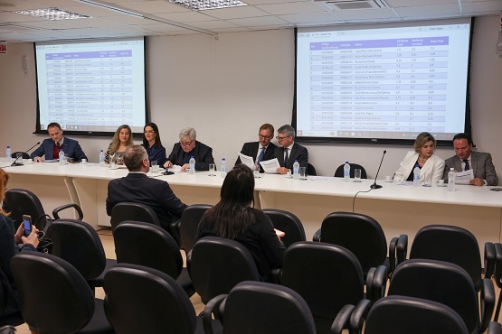A imagem mostra uma sala de conferências com várias pessoas sentadas em uma mesa longa na frente da sala. Há oito pessoas sentadas na mesa, todas vestidas formalmente. Atrás delas, há duas grandes telas de projeção exibindo tabelas com informações. Na frente da mesa, há várias cadeiras dispostas em fileiras, com algumas pessoas sentadas nelas, aparentemente assistindo à apresentação ou reunião. A sala tem iluminação de teto e parece ser um ambiente corporativo ou governamental.