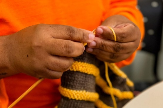A imagem mostra uma pessoa tricotando. Suas mãos estão segurando uma agulha de tricô e um fio de lã amarelo. A peça que está sendo tricotada tem listras amarelas e marrons. A pessoa está vestindo uma blusa laranja. O fundo da imagem está desfocado, mas parece ser um ambiente interno.