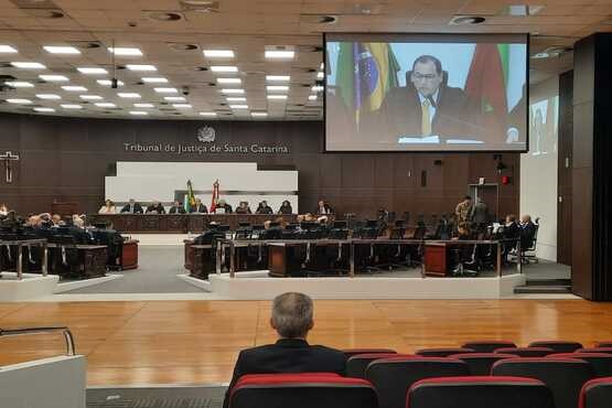 A imagem mostra uma sala de tribunal com várias pessoas sentadas em cadeiras, algumas delas em uma mesa grande na frente da sala. Na parede ao fundo, há um brasão e a inscrição "Tribunal de Justiça de Santa Catarina". À direita, há uma grande tela de projeção exibindo a imagem de um homem falando.