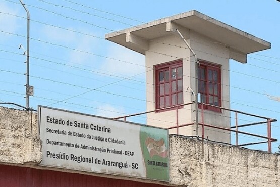 A imagem mostra uma parte do Presídio Regional de Araranguá, localizado no estado de Santa Catarina. Na parte inferior da imagem, há uma placa que diz: "Estado de Santa Catarina, Secretaria de Estado da Justiça e Cidadania, Departamento de Administração Prisional - DEAP, Presídio Regional de Araranguá - SC". Acima da placa, há uma torre de vigilância com janelas de vidro e uma estrutura de concreto. A torre está cercada por arame farpado e há um céu azul ao fundo.