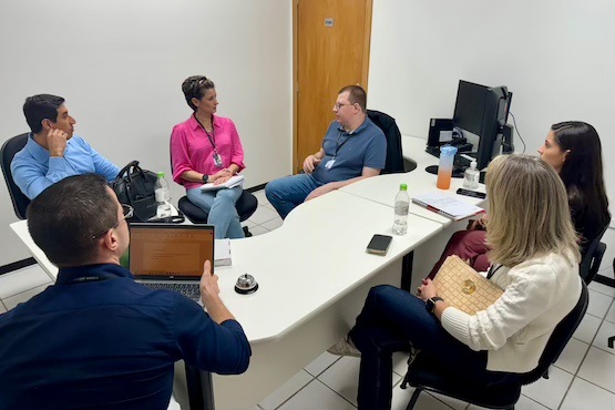 A imagem mostra seis pessoas sentadas em uma sala de reunião. A sala é pequena e tem paredes brancas. Há uma mesa branca em formato de "L" no centro da sala.   - No lado esquerdo da mesa, há um homem de camisa azul claro, com a mão no queixo, parecendo pensativo. - Ao lado dele, uma mulher de camisa rosa está falando, gesticulando com as mãos. - No centro da mesa, um homem de camisa azul escura está de costas para a câmera, usando um laptop. - Do outro lado da mesa, um homem de camisa azul escura está sentado, conversando com a mulher de camisa rosa. - À direita, uma mulher de cabelos loiros e blusa branca segura uma bolsa de palha no colo. - Ao lado dela, uma mulher de cabelos castanhos e blusa preta.  Na mesa, há garrafas de água, um celular e alguns papéis. No fundo da sala, há uma porta de madeira e um computador com dois monitores.